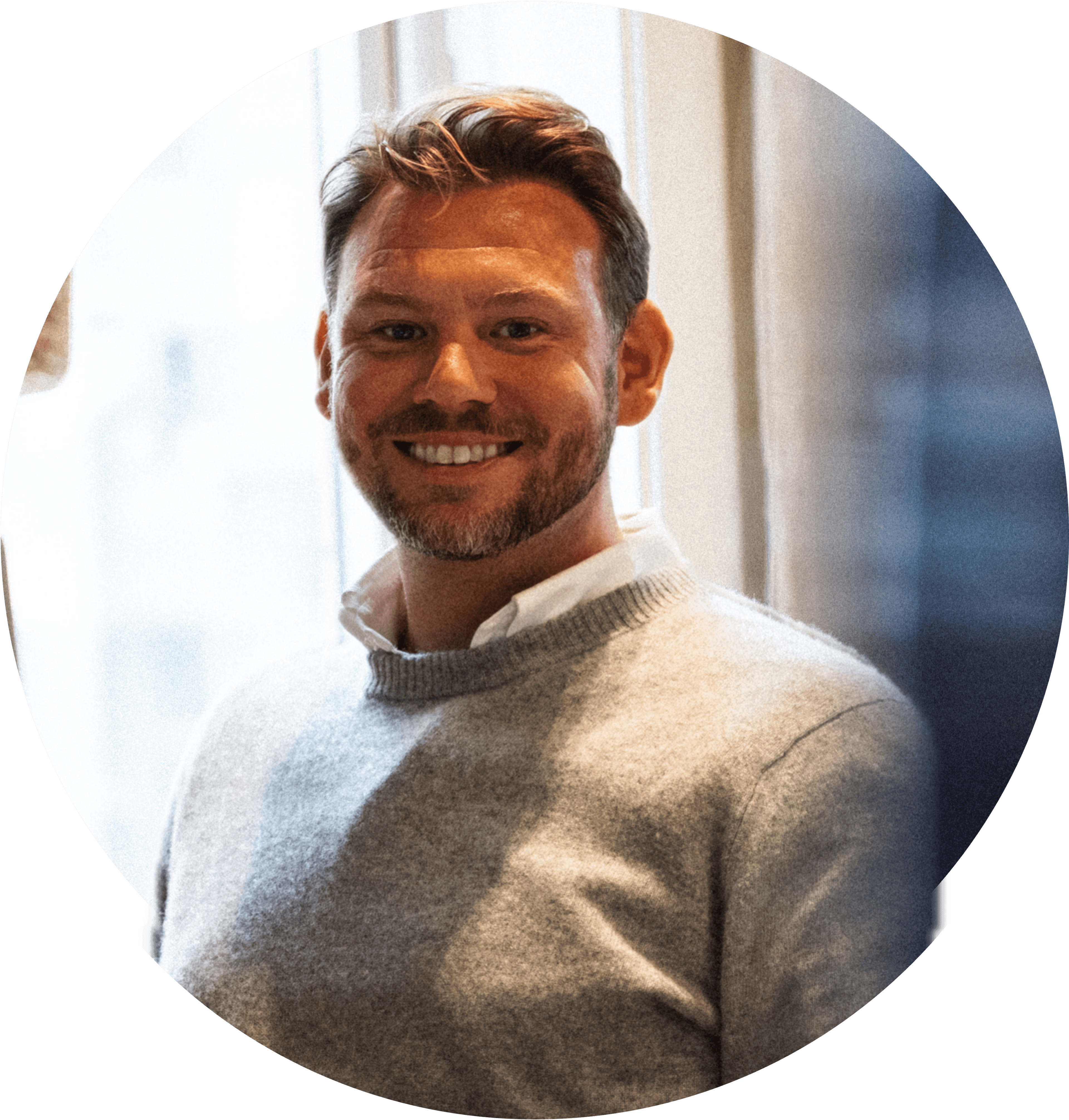 Daniel smiler bredt, iført en grå trøje, med kort brunt hår og lidt skæg. Foto.